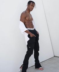 A shirtless man in black pants posing against a white wall.